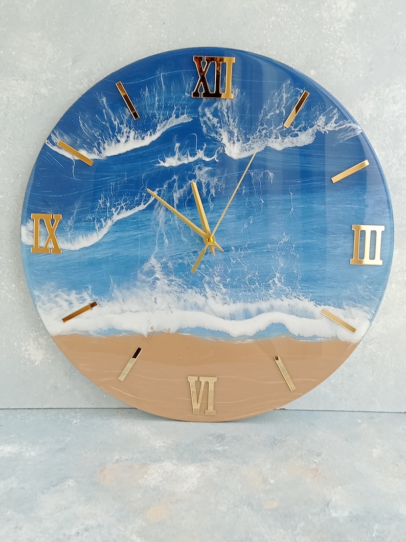 Puede incluir: Reloj de pared redondo con tem&aacute;tica de playa. La esfera del reloj presenta un dise&ntilde;o de oc&eacute;ano azul con olas blancas y una playa beige en la parte inferior. Los n&uacute;meros romanos y las manecillas doradas completan el dise&ntilde;o.
