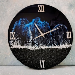 May include: Round wall clock with a black background and a blue and white wave design. The clock face features silver Roman numerals and hands. The bottom of the clock has a textured black surface.