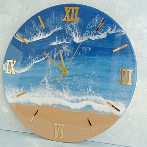 Puede incluir: Reloj de pared redondo con dise&ntilde;o de playa. La esfera del reloj presenta una escena de oc&eacute;ano azul y blanco con n&uacute;meros romanos y manecillas doradas. La parte inferior del reloj es de color beige, asemej&aacute;ndose a arena.