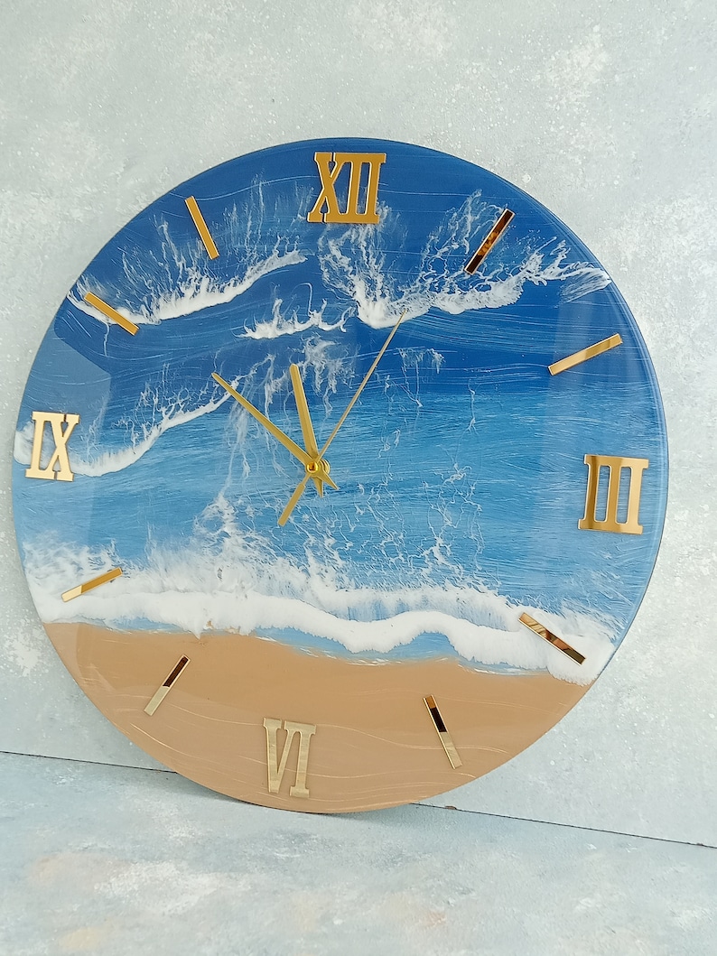 Puede incluir: Reloj de pared redondo con dise&ntilde;o de playa. La esfera del reloj presenta una escena de oc&eacute;ano azul y blanco con n&uacute;meros romanos y manecillas doradas. La parte inferior del reloj es de color beige, asemej&aacute;ndose a arena.