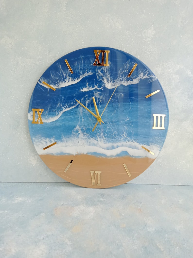 Puede incluir: Reloj de pared redondo con dise&ntilde;o de escena de playa. La esfera del reloj presenta un oc&eacute;ano azul con olas blancas y una playa arenosa. Los n&uacute;meros romanos y las manecillas doradas completan el dise&ntilde;o.