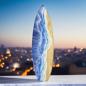 May include: A wooden surfboard with a blue and white resin wave design. The surfboard is standing upright on a white surface.