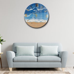 Puede incluir: Reloj de pared redondo con dise&ntilde;o de escena de playa. La esfera del reloj presenta un oc&eacute;ano azul, olas blancas y una orilla arenosa. Los n&uacute;meros romanos dorados marcan las horas. El reloj est&aacute; montado en una pared blanca.