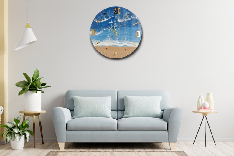 Puede incluir: Reloj de pared redondo con dise&ntilde;o de escena de playa. La esfera del reloj presenta un oc&eacute;ano azul, olas blancas y una orilla arenosa. Los n&uacute;meros romanos dorados marcan las horas. El reloj est&aacute; montado en una pared blanca.