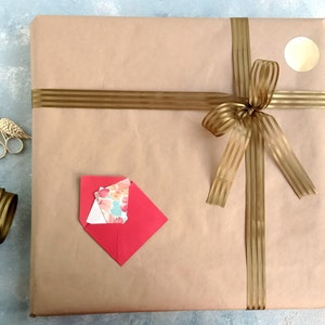 Puede incluir: Un paquete envuelto en papel marr&oacute;n, atado con una cinta y un lazo dorados. Un sobre rojo con tarjetas florales, tijeras doradas y un rollo de cinta dorada est&aacute;n junto al paquete. Una pegatina dorada est&aacute; en el paquete.