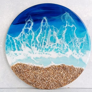 May include: Round resin art piece depicting an ocean scene. The top portion is dark blue, transitioning to turquoise and white, resembling waves. The bottom edge features a strip of small, tan pebbles, creating a beach effect. The artwork is a decorative piece.