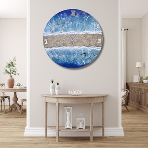 Könnte beinhalten: Eine runde Wanduhr mit einer blauen und weißen Strandszene. Das Zifferblatt der Uhr hat römische Ziffern und einen weißen Hintergrund. Die Uhr hängt an einer weißen Wand.