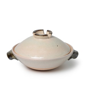 May include: A white ceramic pot with a brown rim and handles. The pot has a lid with a small hole in the center.