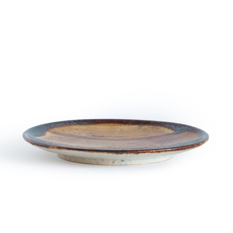 May include: A brown and white ceramic plate with a slightly raised rim. The plate has a rustic, hand-thrown look and is glazed in a brown and white color.