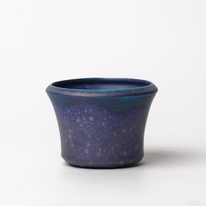 May include: A small, flared ceramic bowl featuring a deep violet and blue glaze with a speckled, mottled texture. The rim is smooth and dark, transitioning into a lighter, dotted pattern across the body of the vessel, which has a flat base.