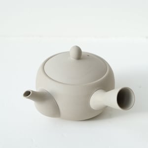 May include: A light grey ceramic teapot with a rounded body and a spout on the side. The teapot has a lid with a small knob on top.