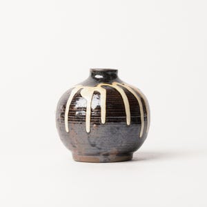 May include: A small, round ceramic vase with a dark brown and charcoal gray glazed finish. The surface features horizontal ridges and a decorative cream-colored glaze that drips vertically from the narrow neck down the sides of the vessel.
