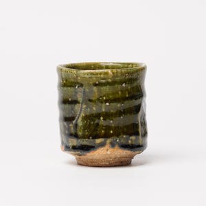 May include: A handcrafted ceramic cup with a textured, dark green glaze. The cup has a rough, organic shape and a natural, unglazed base. The glaze features subtle variations in colour and texture, giving it a unique, artisanal look.