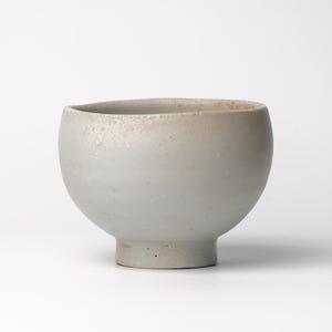 May include: A handcrafted, light gray ceramic bowl with a rounded body and a small, sturdy base. The rim has a subtle, light brown gradient. The bowl's surface has a slightly textured appearance, suggesting a natural, artisanal quality. This bowl is perfect for serving soups or snacks.