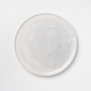 May include: A round, white ceramic plate with a delicate, web-like pattern in a lighter shade of white. The design is centered and resembles a network of fine lines and cracks, creating a subtle, textured effect. The plate is on a white background.