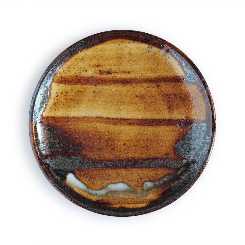 May include: A brown ceramic plate with a striped glaze pattern. The plate has a dark brown rim and a light brown center with a white glaze in the center.
