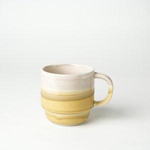 May include: A ceramic mug with a layered design in cream, tan, and yellow. The mug has a rounded handle and a stacked appearance. The mug is suitable for hot or cold drinks. The mug's dimensions are approximately 10 cm in height.