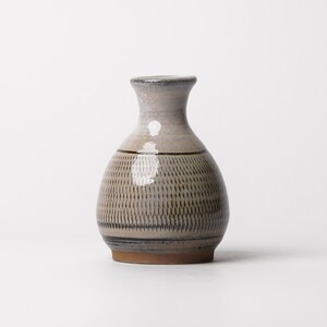 May include: A ceramic sake bottle with a rounded body and a narrow neck. The exterior features a light grey glaze with a dark brown horizontal band and a repeating pattern of vertical dashes. The base is unglazed, showing the natural brown clay.