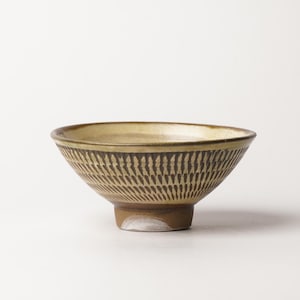 May include: A ceramic bowl featuring a wide, flared rim and a tapered base. The exterior is decorated with a repeating pattern of dark brown, vertical, dash-like carvings against a light tan background. The interior has a smooth, cream-colored glaze.