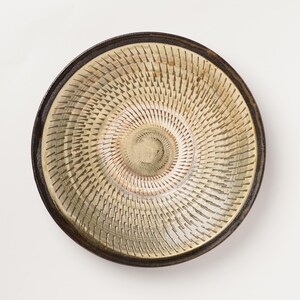 May include: A circular ceramic bowl with a dark brown rim and a light beige interior. The interior surface features a repeating pattern of small, incised vertical marks arranged in a spiral formation radiating from the center of the bowl.