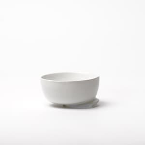 May include: A white ceramic bowl with a rounded shape, resting on a small, light gray base. The bowl has a smooth, glossy finish and a simple, modern design. The bowl is likely used for serving food or as a decorative item.