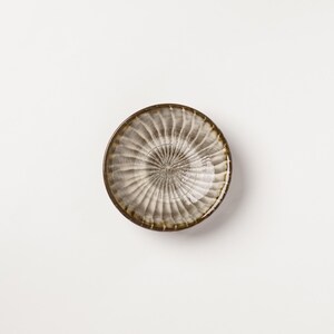 May include: A small, round ceramic bowl featuring a radial, fluted pattern that radiates from the center. The surface displays a variegated glaze with tones of grey, brown, and cream, creating a textured appearance with a dark, defined rim.