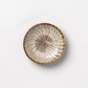 May include: A small, round bowl featuring a swirling, radial pattern in shades of beige, cream, and dark brown. The interior surface displays a textured, ribbed design that radiates from the center toward the smooth, circular rim of the vessel.