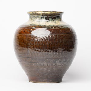 Puede incluir: Un jarrón de cerámica marrón con una superficie texturizada y patrones decorativos de ondas y triángulos. El jarrón tiene un cuerpo ancho y un cuello más estrecho con un borde crema y verde. El jarrón mide unos 30 cm de alto.