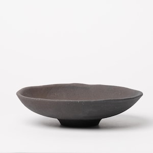 May include: A small, dark gray ceramic bowl with a shallow, irregular rim. The bowl has a rounded base and a slightly textured surface. The overall shape is organic and minimalist, suitable for serving small portions or as a decorative piece.
