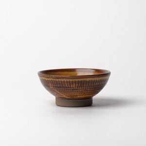 May include: A small ceramic bowl with a rounded shape and a raised footed base. The exterior features a textured, stamped pattern in shades of brown and ochre, while the interior has a smooth, glossy finish in a warm, golden-brown glaze.