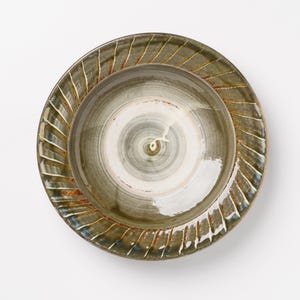 May include: A round ceramic bowl with a wide, flat rim featuring a series of diagonal, carved grooves. The surface displays a variegated glaze in shades of olive green, brown, and cream, with visible circular brushwork patterns in the center.