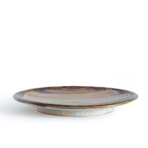 May include: A brown and white ceramic plate with a slightly raised rim. The plate has a rustic, hand-thrown look and is glazed in a brown and white color.