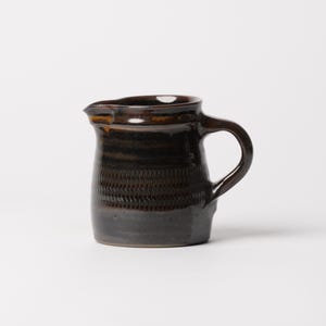 May include: A small ceramic creamer pitcher with a dark brown, glossy glaze. The body features a textured, horizontal band of repeating vertical indentations around the center, a wide rim, and a curved handle on the right side of the vessel.