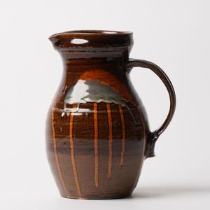 May include: A handmade brown ceramic pitcher with a curved handle and a pouring spout. The pitcher features vertical orange glaze streaks and a glossy finish. The pitcher is approximately 9 inches tall.