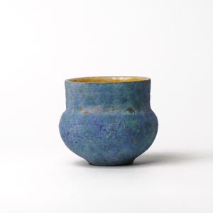 May include: A small, rounded ceramic cup with a textured, mottled surface in shades of deep blue, cobalt, and hints of green. The exterior has a rough, matte finish, while the interior rim reveals a contrasting smooth, warm yellow glaze.