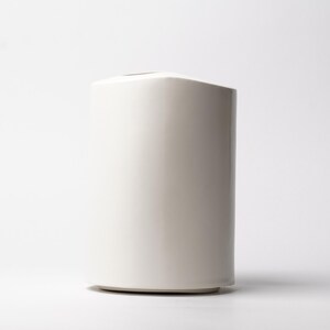 May include: A tall, off-white ceramic vase with a cylindrical shape and a slightly curved top. The vase has a smooth, glossy finish and sits on a small, circular base. The design is minimalist and modern, suitable for various home decor styles.