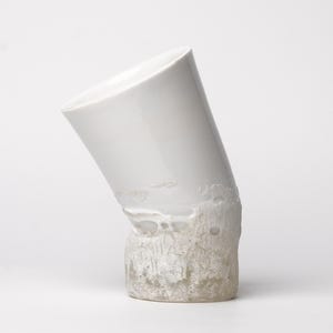 May include: A white ceramic vase with a unique, angled design. The top portion is smooth and glossy, while the base has a textured, rough appearance. The vase is a modern art piece.