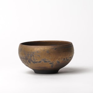 May include: A round ceramic bowl with a metallic bronze and dark brown finish. The exterior features a textured, mottled appearance with darker, uneven patches near the base and a smooth, curved rim. The bowl has a deep, wide basin.