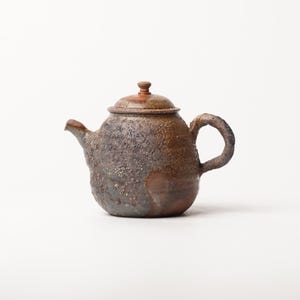 Puede incluir: Una tetera de cerámica hecha a mano con una superficie rugosa y texturizada en tonos marrón oscuro, gris carbón y ocre terroso. La tetera presenta un cuerpo redondeado, un asa curva, un pico corto y una tapa a juego con un pequeño pomo.