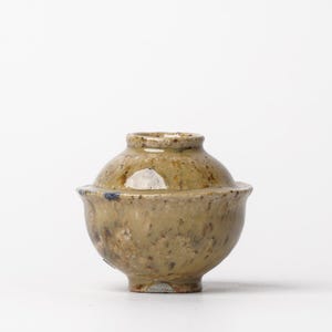 May include: A small, round, ceramic pot with a lid. The pot is glazed in a light brown color with darker brown speckles. The lid has a slightly raised rim.