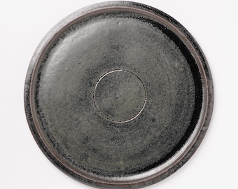 May include: A round, dark gray ceramic plate with a slightly raised rim. The plate has a matte finish and a circular indentation in the center. The plate's color is a blend of dark gray and hints of brown.
