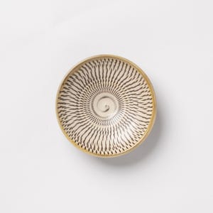 May include: A round ceramic bowl featuring a cream-colored interior decorated with a dark brown radial pattern of small, elongated, dash-like marks that spiral outward from the center toward the smooth, light brown rim of the vessel.