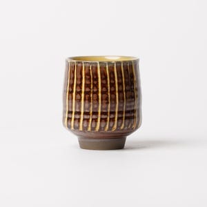 May include: A ceramic cup featuring a cylindrical shape with a rounded base. The exterior displays a dark brown glaze with vertical cream-colored stripes and a textured, grid-like pattern. The interior rim shows a smooth, light yellow glaze finish.