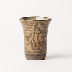 May include: A ceramic tumbler with a flared rim and a tapered base. The exterior features a textured, repeating vertical pattern in shades of mustard yellow and dark brown, with a smooth, light-colored glaze visible along the inner rim.