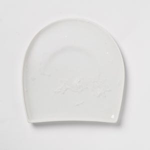 May include: A white, semi-circular plate with a slightly textured surface. The plate has a rounded top and a curved bottom edge. The overall shape is reminiscent of a shallow bowl or serving dish. The plate is set against a plain white background.