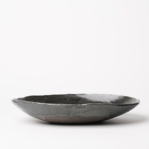 May include: A handmade, dark gray ceramic bowl with a rough, uneven rim. The bowl has a shallow, oval shape and a dark brown base. The surface has a slightly speckled appearance, suggesting a unique glaze.