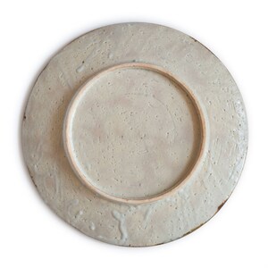 May include: The underside of a ceramic plate with a light gray glaze and a textured surface. The plate has a raised rim and a smooth, unglazed center.