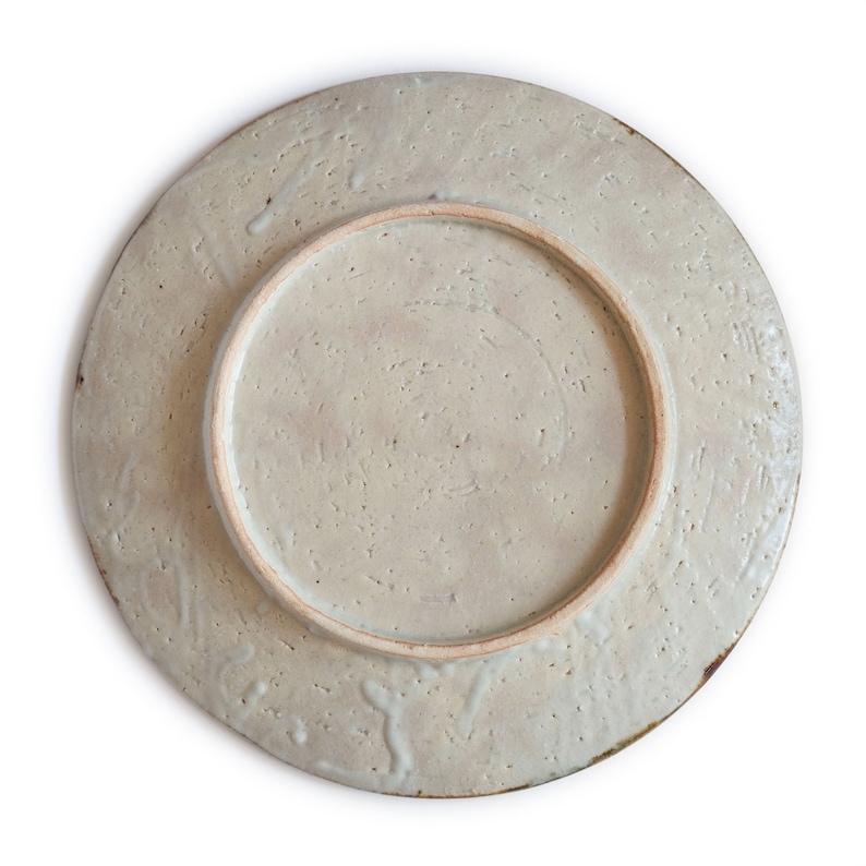 May include: The underside of a ceramic plate with a light gray glaze and a textured surface. The plate has a raised rim and a smooth, unglazed center.