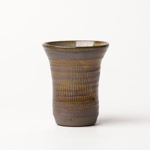May include: A cylindrical ceramic tumbler with a flared rim, featuring a dark brown base glaze overlaid with a textured, vertical pattern of lighter golden-brown markings that wrap around the exterior surface of the vessel.