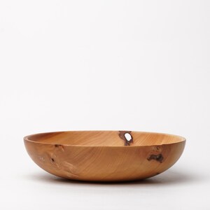 Könnte beinhalten: Eine handgefertigte Holzschale mit natürlicher, hellbrauner Oberfläche. Die Schale hat ein breites, flaches Design und sichtbare Holzmaserung. Sie weist einen Knoten und ein kleines Loch auf, was ihren rustikalen Charme unterstreicht. Ideal zum Servieren oder Ausstellen.